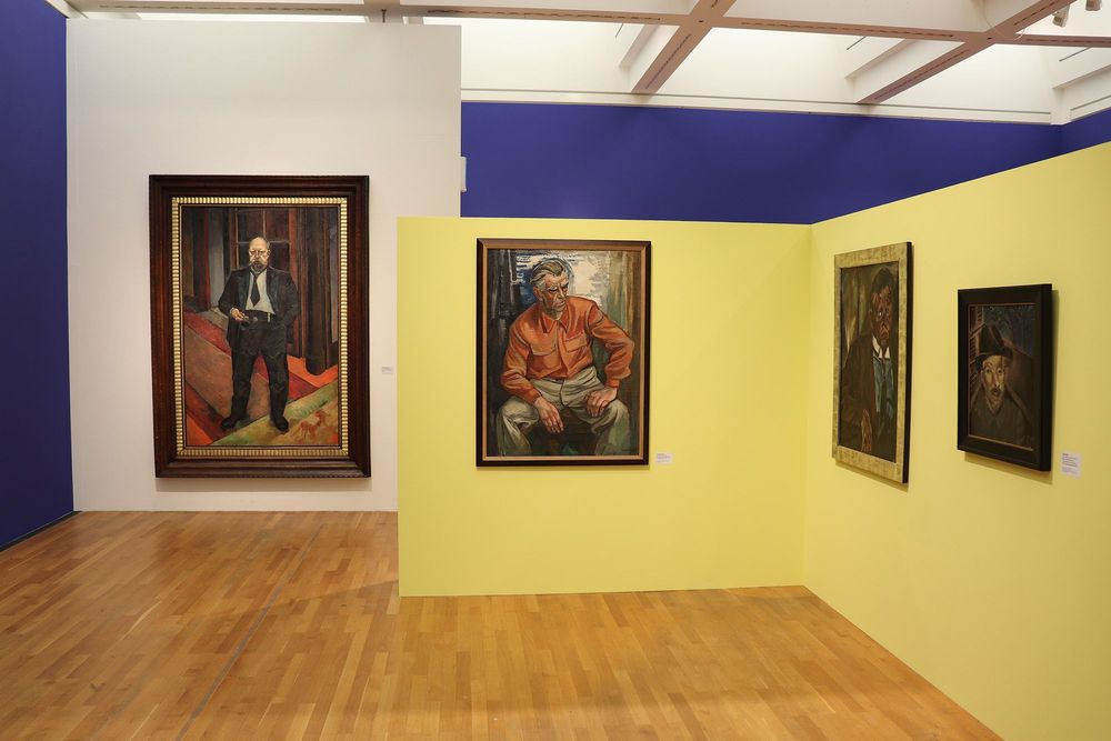 Die Ausstellungsansicht zeigt auf der rechten Seite eine gelbe Wand, an der drei Gemälde hängen. Links an der blau-weißen Wand hängt ein weiteres Gemälde, welches einen Mann im schwarzen Anzug und mit Brille in der Hand zeigt.