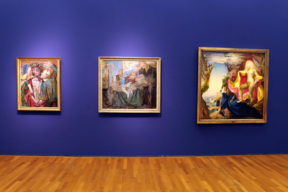 An einer blauen Wand hängen drei große Gemälde. 