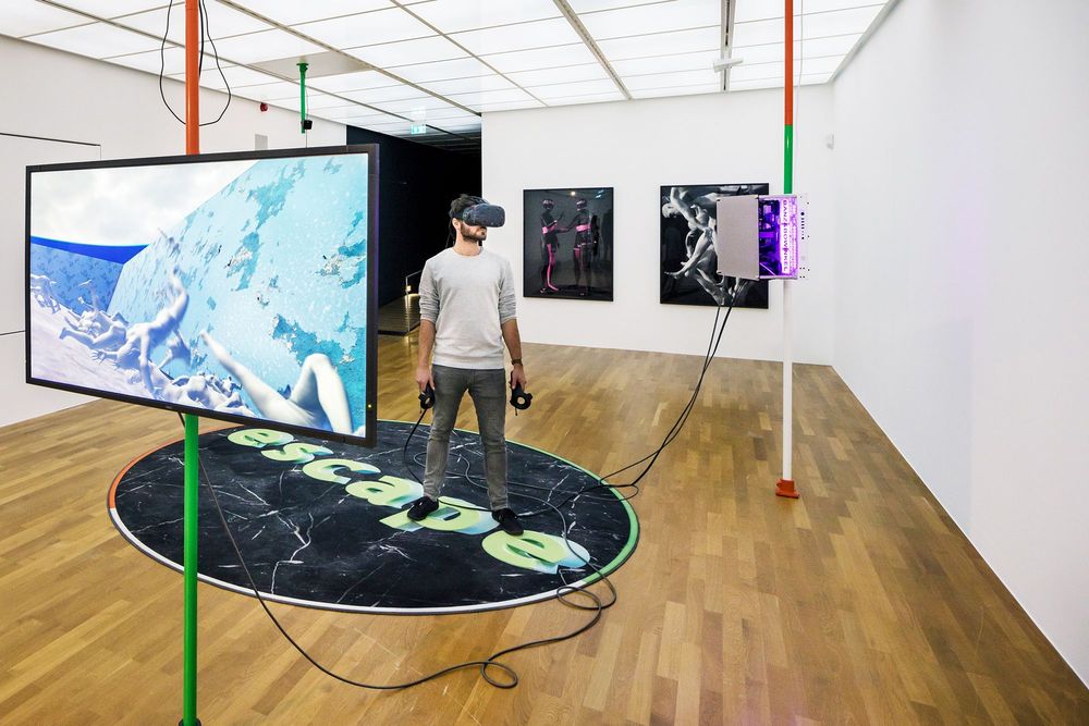 Die Ausstellungsansicht zeigt einen Mann der eine VR-Brille trägt und auf einem runden Teppich mit dem Schriftzug "escape" steht. Links im Vordergrund befindet sich ein Bildschirm auf dem ein Video abspielt.