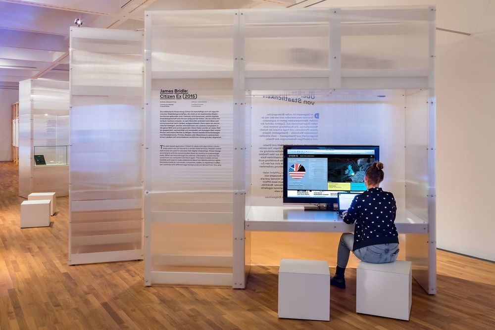 Die Ausstellungsansicht zeigt eine Stellwand mit Ausstellungstexten und einem Tisch, auf dem sich ein Laptop und ein großer Bildschirm befinden. Eine Frau sitzt vor dem Laptop.