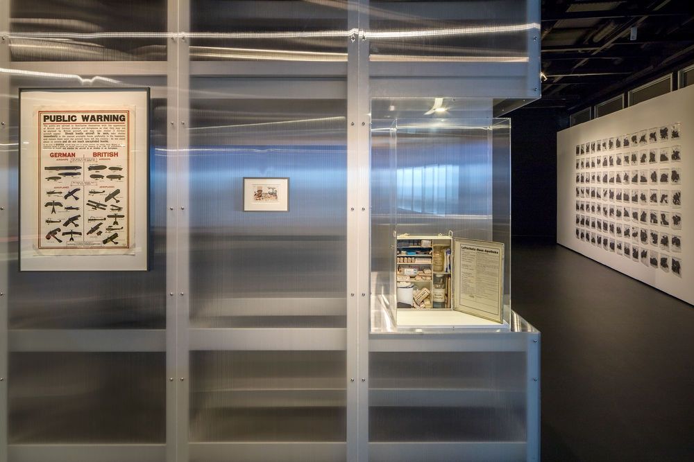 Die Ausstellungsansicht zeigt eine Ausstellungswand an der verschiedene Bilder angebracht sind, sowie ein Schaukasten in dem sich ein  Luftschutz Apothekenkasten befindet.