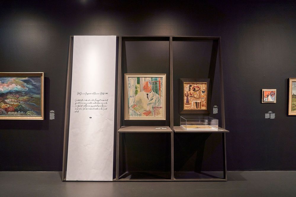 Die Ausstellungsansicht zeigt eine Stellwand mit Ablageflächen, auf denen ein altes Buch in einem Glaskasten liegt. An der Wand hängen verschiedene Gemälde.