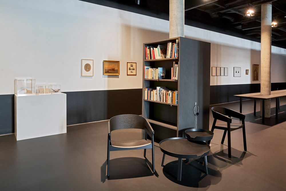 Die Ausstellungsansicht zeigt ein großes Regal in dem viele Bücher stehen. Davor befinden sich ein kleiner Tisch und Stühle. An der Wand stehen auf einem weißen Podest mehrere Exponate.