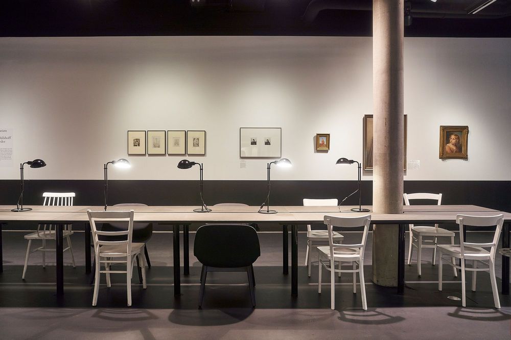 Die Ausstellungsansicht zeigt mehrere aneinandergereihte Tische, auf denen Leselampen stehen. An der Wand im Hintergrund hängen verschiedene Bilder.