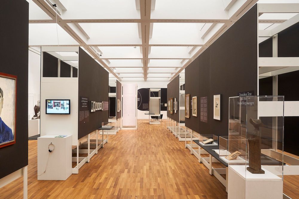 Die Ausstellungsansicht zeigt einen Flur der aus aufgestellten Wänden besteht. An den Wänden hängen Bilder und Texte, sowie Objekte in Glaskästen. 