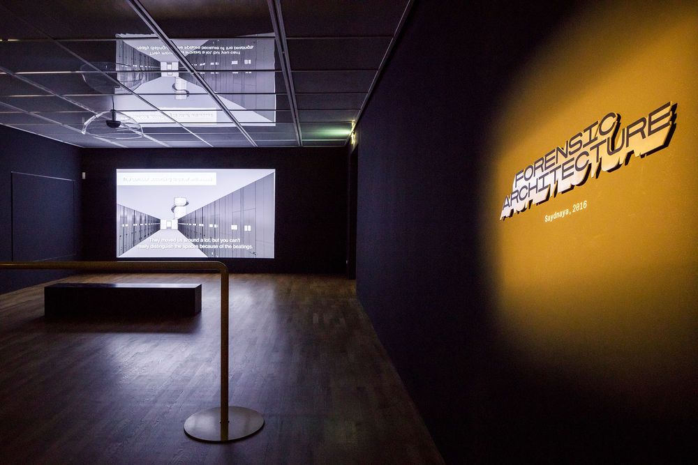 Die Ausstellungsansicht zeigt einen Raum mit schwarzen Wänden in dem sich eine Sitzbank und eine Leinwand befinden, auf welche ein Video projiziert wird. Rechts im Vordergrund hängt ein Text an der Wand, der mit gelb, orangenem Licht beleuchtet wird.