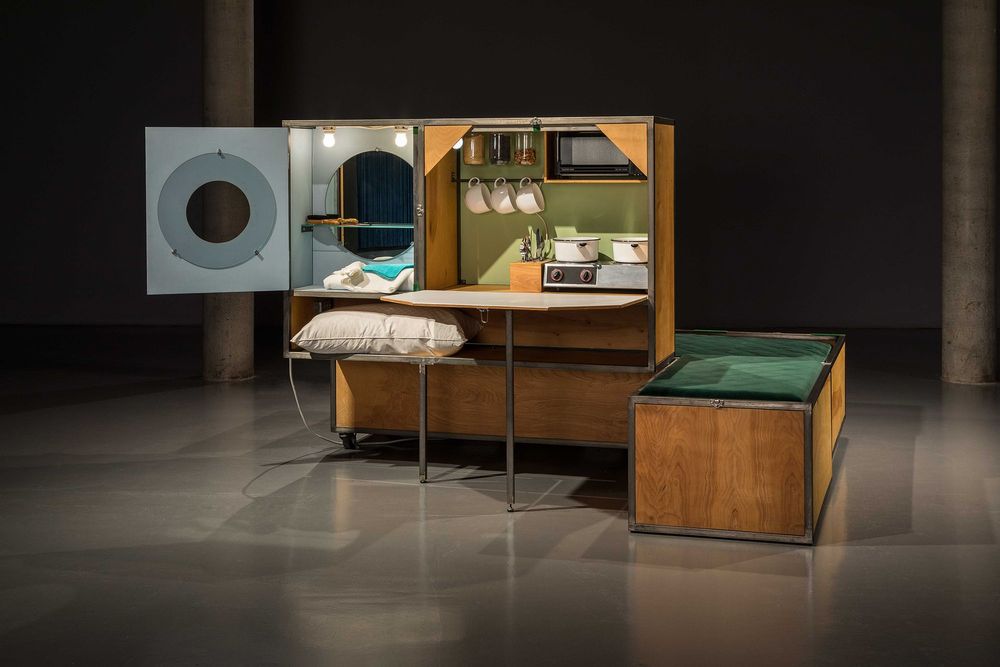 Die Ausstellungsansicht zeigt eine mobile Sitzbank aus Holz mit dunkelgrünen Stoffbezügen. An der Sitzbank ist eine Kochnische, sowie ein aufklappbarer Tisch und daneben ein aufklappbarer Badezimmerspiegel mit Waschbecken und Handtüchern befestigt.