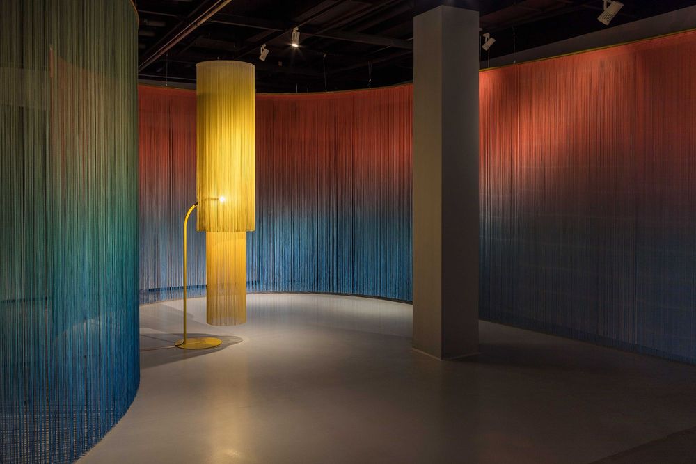 Die Ausstellungsansicht zeigt einen mehrfarbige, durchsichtigen Vorhang der den Raum trennt, darin steht eine gelbe Lampe, die von einem weiteren gelben Vorhang umgeben ist.