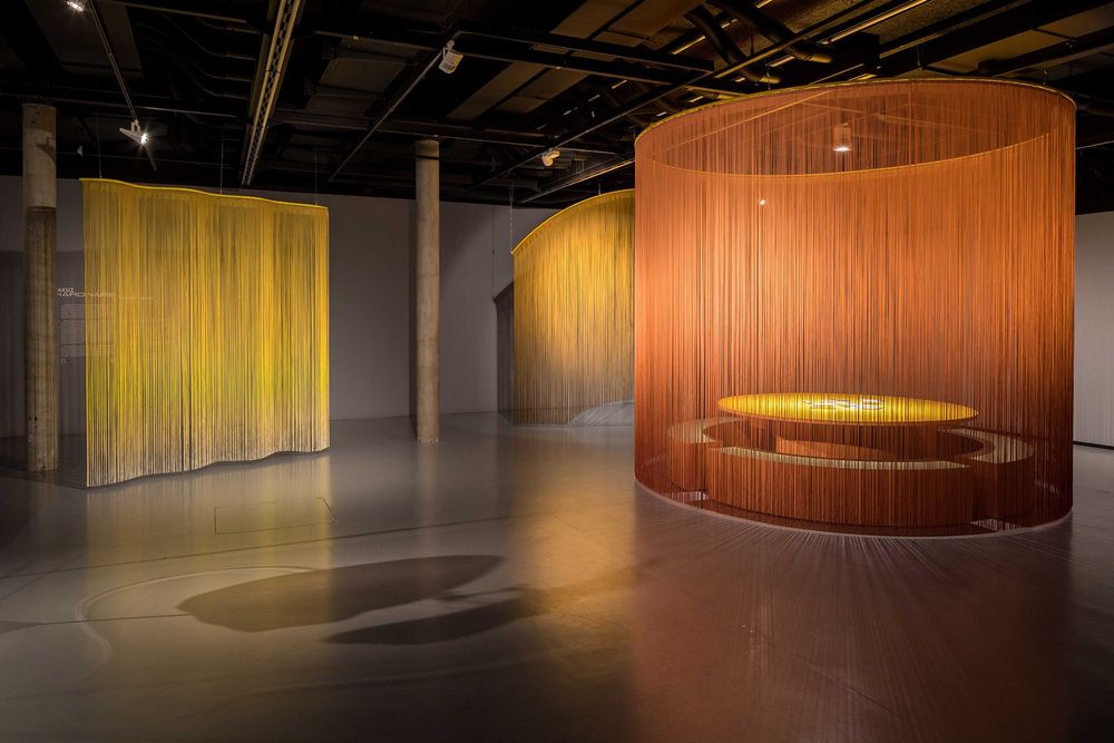 Die Ausstellungsansicht zeigt einen grauen Raum in dem mehrere kleine Räume durch farbige, durchsichtige Vorhänge abgetrennt sind. Hinter einem kreisförmigen Vorhang befindet sich ein runder Tisch mit Sitzbänken.