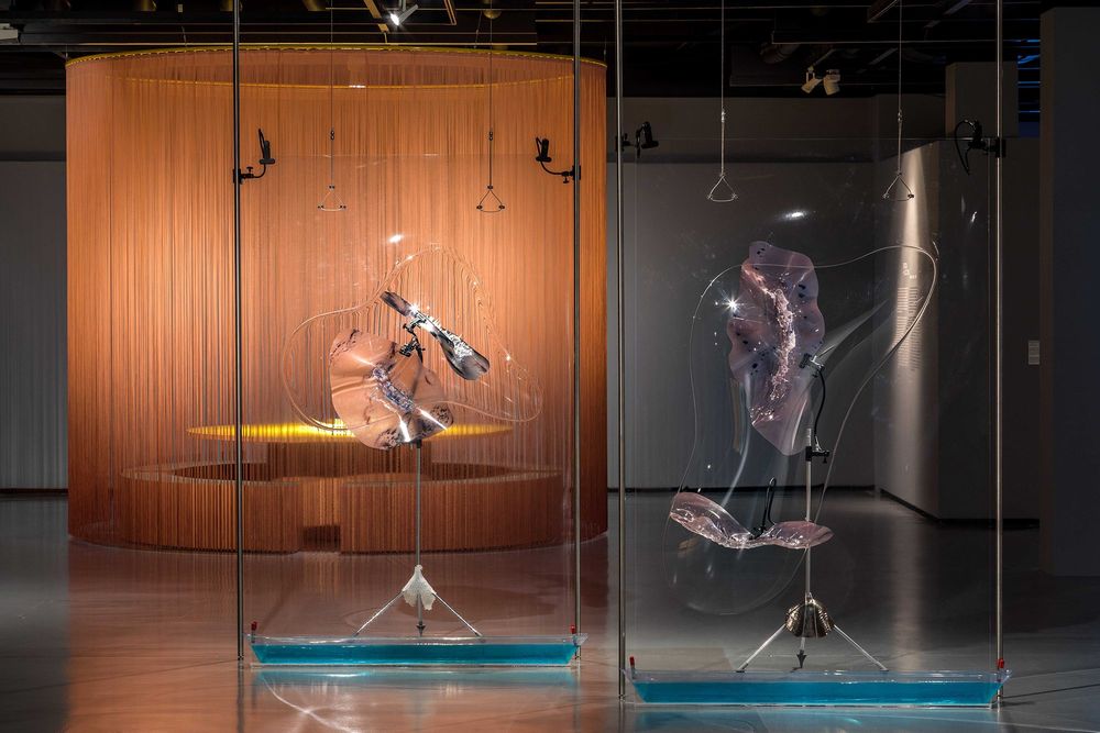 Die Ausstellung zeigt im Vordergrund zwei Installationen, dahinter befindet sich einkreisförmigen Vorhang hinter dem ein runder Tisch mit Sitzbänken steht.