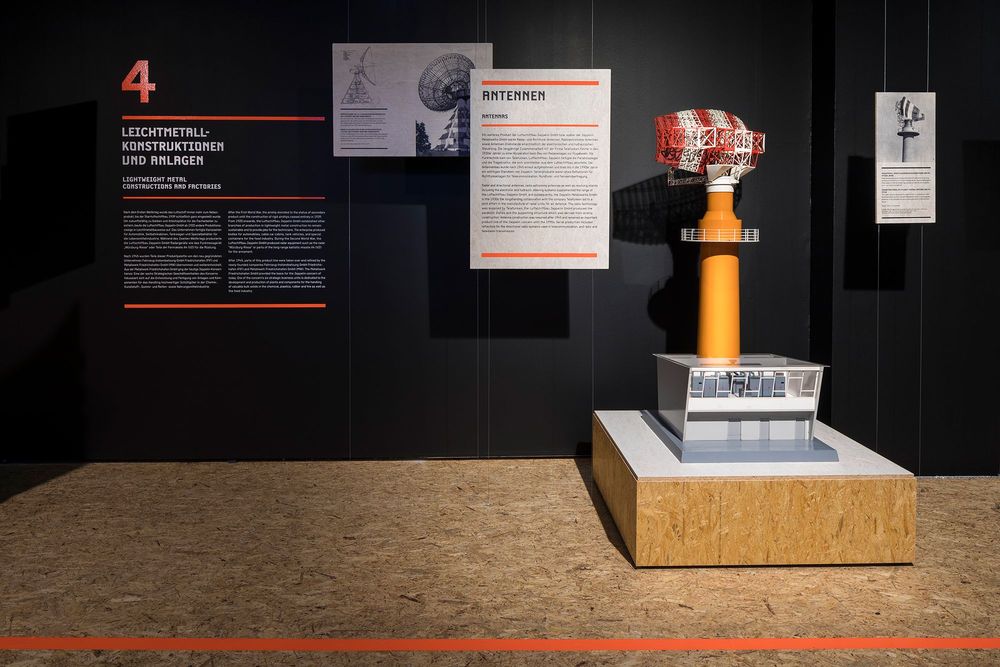 Die Ausstellungsansicht zeigt Bildmaterial und Ausstellungstexte, die an einer schwarzen Wand befestigt sind. Auf einem Podest steht das Modell einer großen Antenne.