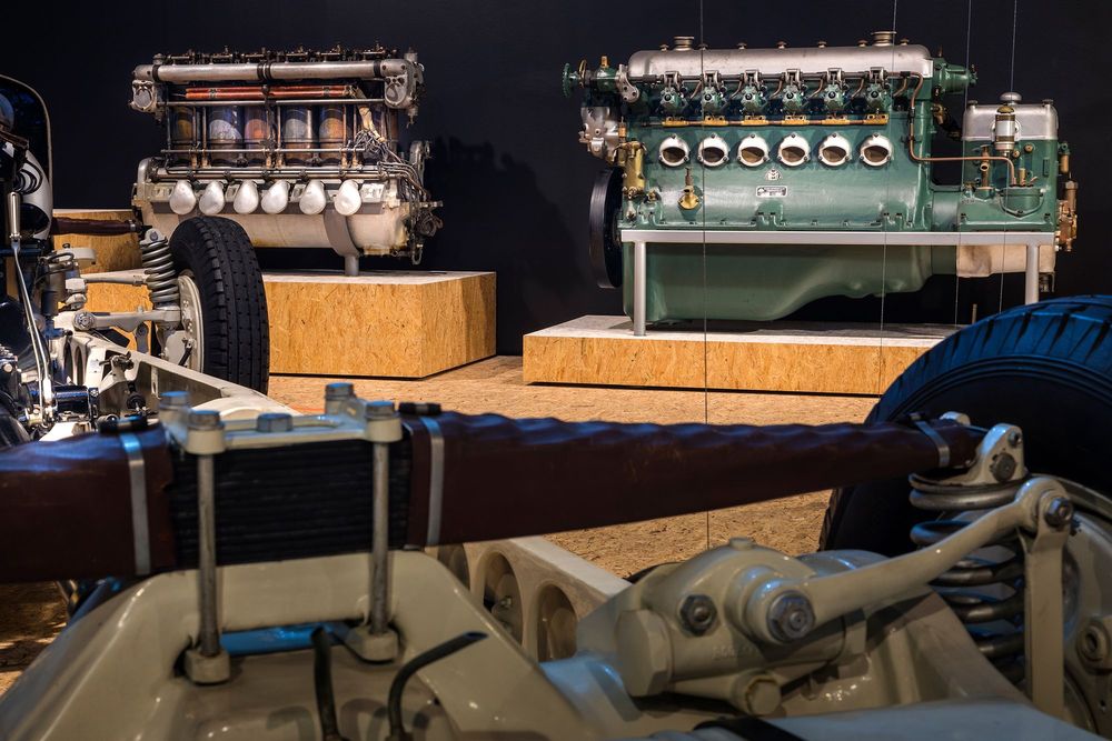 Die Ausstellungsansicht zeigt zwei große Antriebsmotoren, die auf Podesten stehen. Im vorderen Bereich der Aufnahme ist ein Automobil-Modell zu sehen.