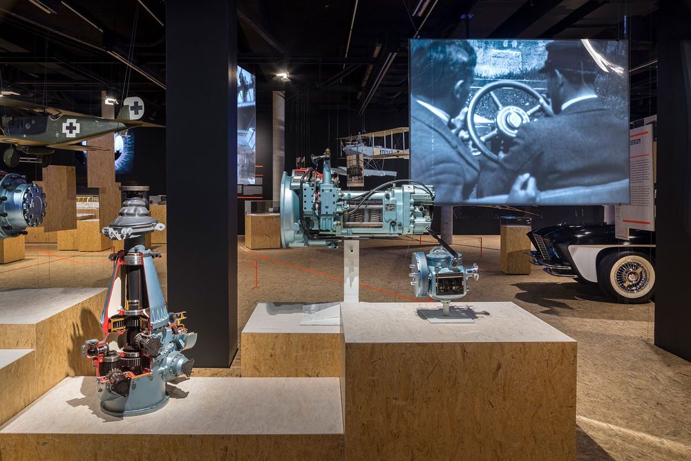 Die Ausstellungsansicht zeigt diverse Ausstellungsobjekte, die in unterschiedlichen Höhen auf Podesten platziert sind. Rechts in der Aufnahme hängt ein großes Bild von der Decke, welches zwei Männer die hinter einem Lenkrad sitzen abbildet.
