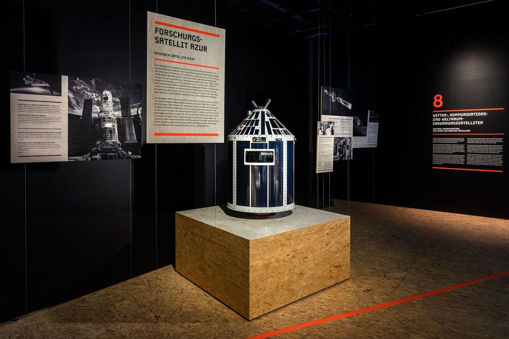 Die Ausstellungsansicht zeigt das Modell eines Forschungssatellit, der auf einem Podest steht. Dahinter an der schwarzen Wand hängen Bilder und Ausstellungstexte.