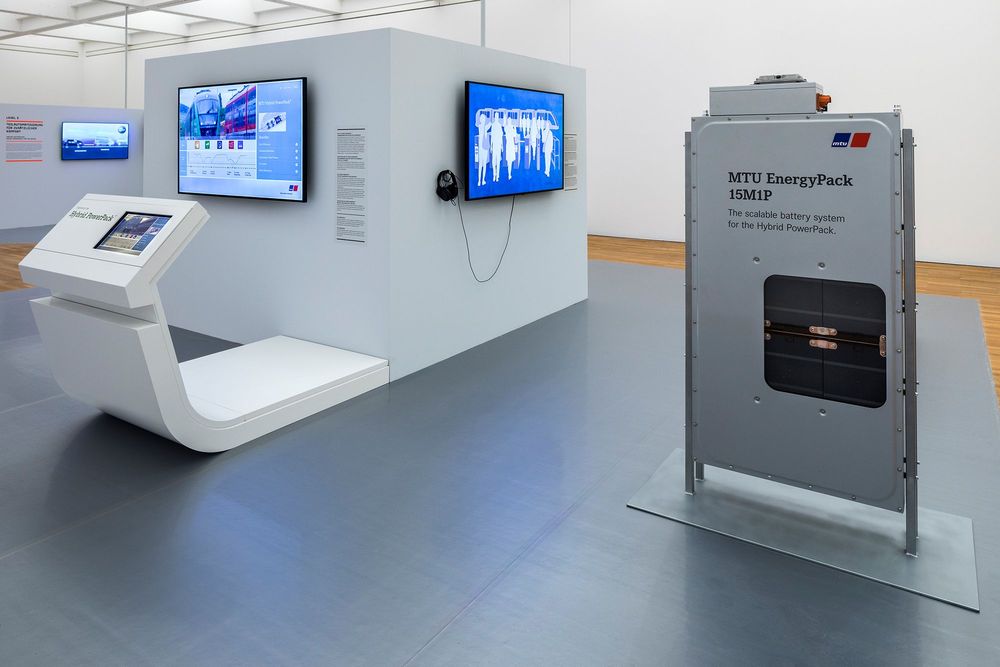 Die Ausstellungsansicht zeigt eine Installation bestehend aus einem weißen großen Würfel, an dem zwei Bildschirme und Texte hängen. Daneben im Vordergrund steht ein Art Batteriekasten. 