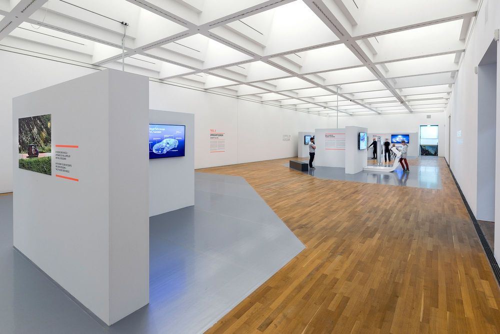Die Ausstellungsansicht zeigt einen weißen Raum in dem kleinere Wände mit Texten und Bildschirmen aufgebaut sind. Besucher*innen befinden sich an Stationen oder lesen Ausstellungstexte.