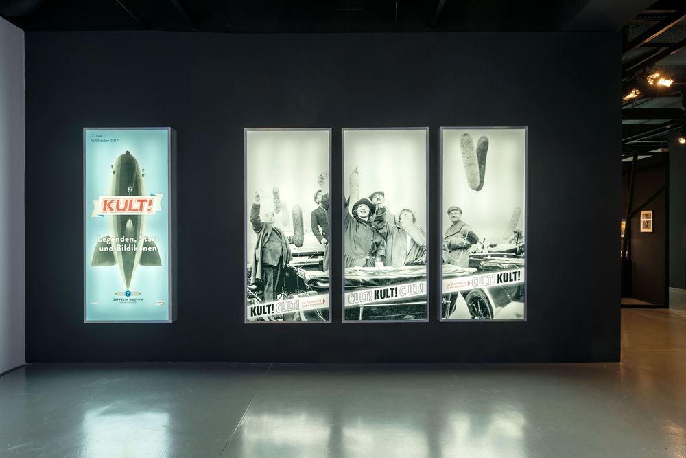 Die Ausstellungsansicht zeigt die Grafik der Ausstellung "KULT!", sowie ein historisches Bild, welches über drei Leuchtkästen verteilt ist. 