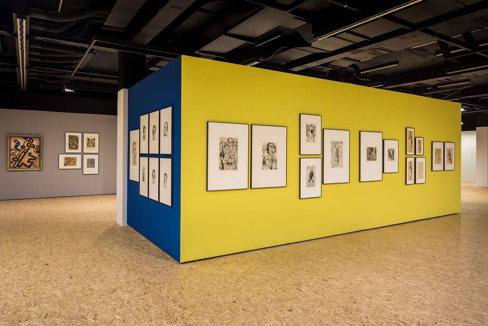 Die Ausstellungsansicht zeigt eine gelbe und eine blaue Wand, die aneinander grenzen. An beiden Wänden hängen verschiedene Zeichnungen von Max Ackermann. Im Hintergrund befindet sich eine graue Wand, an der ebenfalls fünf Bilder hängen.