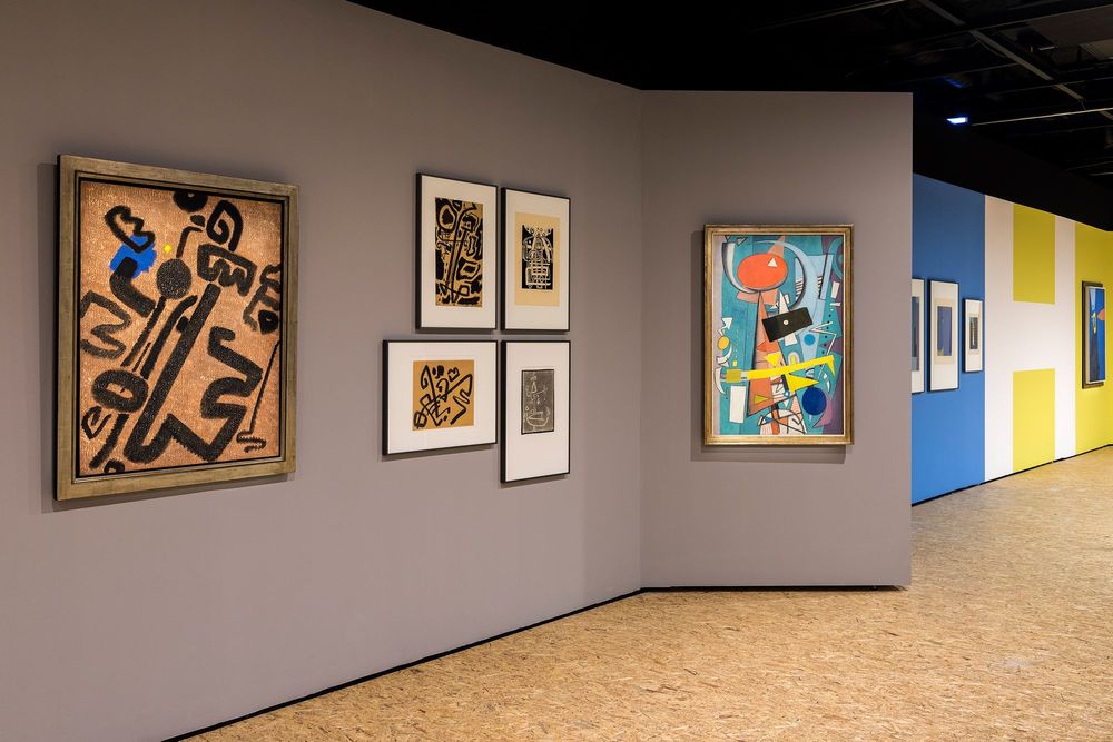 Die Ausstellungsansicht zeigt im Vordergrund eine graue Wand an der sechs Bilder hängen, zwei davon sind farbenfroh und bunt. Hinter der grauen Wand grenzen eine blaue und eine gelbe Wand an, an denen ebenfalls Bilder hängen.