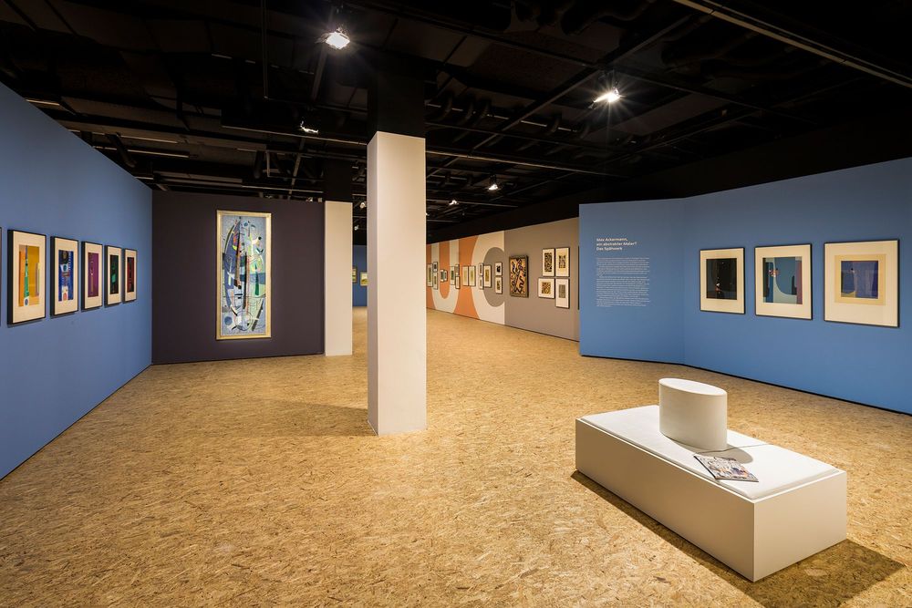 Die Ausstellungsansicht zeigt einen offenen Raum mit einer weißen Sitzbank für Besucher*innen. An den blauen Wänden im Vordergrund, sowie den Wänden im Hintergrund hängen verschiedene Gemälde von Max Ackermann.