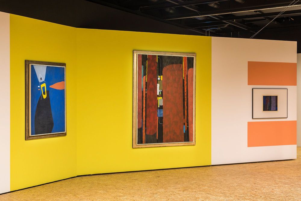 Die Ausstellungsansicht zeigt eine weiß-gelbe Wand, an der zwei große bunte Gemälde, sowie ein kleineres Gemälde von Max Ackermann hängen. 