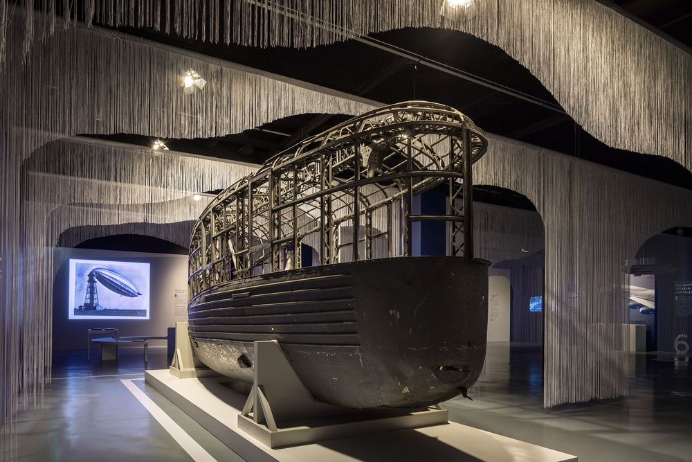 Die Ausstellungsansicht zeigt die Gondel des Luftschiffs L30, die auf zwei Stützen liegt. Darüber hängen von der Decke verkürzte weiße Vorhänge aus Schnürren.