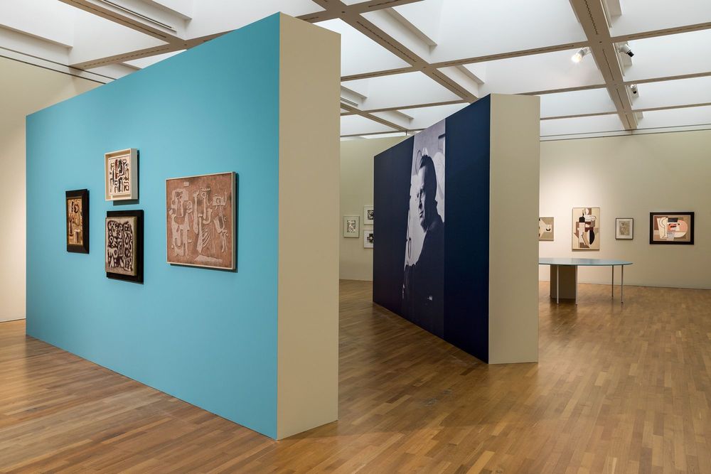 Die Ausstellungsansicht zeigt zwei breite Wände,  die im Raum aufgestellt sind. An der vorderen Wand hängen vier Gemälde, an der hinteren Wand ist ein großes Porträt von Willi Baumeister abgedruckt. Dahinter ist ein runder Tisch zu erkennen.