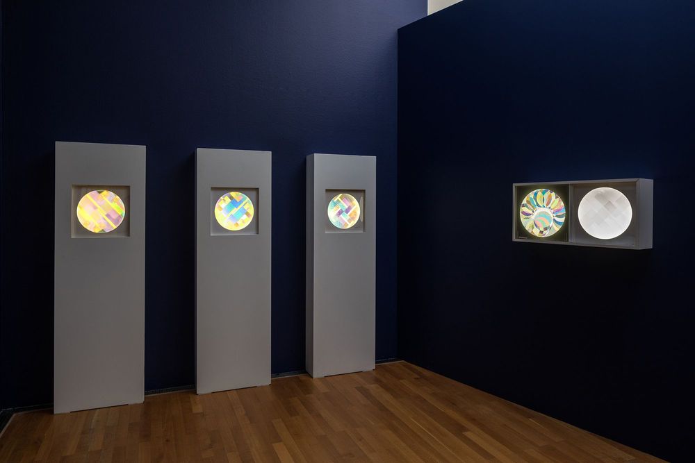 Die Ausstellungsansicht zeigt fünf variochromatische Objekte von Marta Hoepffner. Drei davon befinden sich in weißen Podesten eingerahmt. Vier Objekte sind sehr farbenfroh und leuchten kräftig.