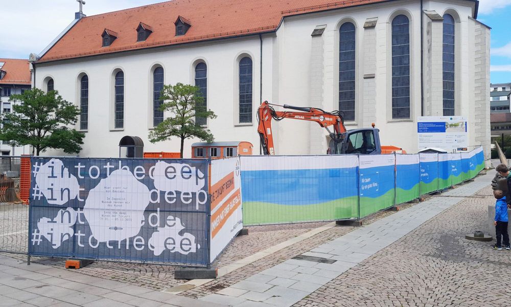 Vor der Kirche in Friedrichshafen ist einer der Bauzäune passend zur Ausstellung "Into the deep. Minen der Zukunft" bedruckt.