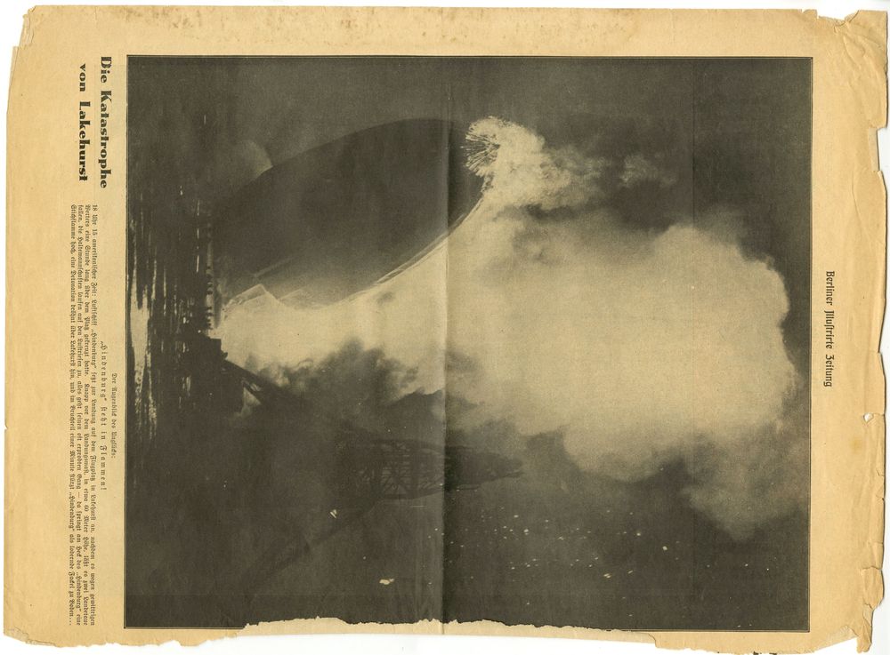 Das Foto zeigt die brennende „Hindenburg“ mit dem Bug nach oben und dem Heck nach unten. Der Ankermast ist als Schatten zu sehen. Eine riesige Rauchwolke steigt auf. Als Schatten erkennt man einige Leute.