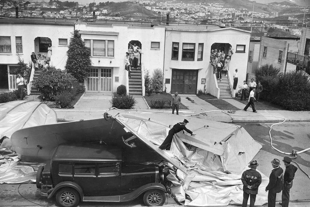 Die historische Aufnahme zeigt ein abgestürztes Luftschiff in einer Wohngegend. Es liegt mitten auf der Straße vor einer Reihe Wohnhäuser. Schaulustige treten aus ihren Häusern und beobachten Feuerwehr und Polizei. 