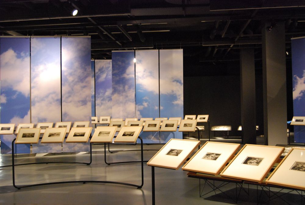 Die Ausstellungsansicht von "Eduard Spelterini Fotografien des Ballonpioniers" zeigt die Aneinanderreihung von Bilderrahmen, die im Raum angeordnet sind. Im Hintergrund stehen Banner, die mit einem Himmel und Wolken Motiv bedruckt sind.