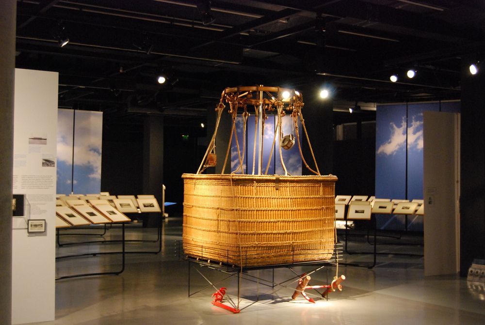 Die Ausstellungsansicht von "Eduard Spelterini Fotografien des Ballonpioniers" zeigt den Korb eines Heißluftballons, der in der Mitte des Raumes steht. Darum herum sind Bilderrahmen nebeneinander angeordnet.