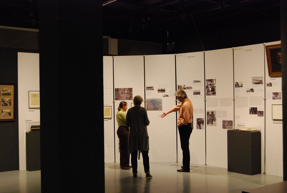 Die Ausstellungsansicht von "Eduard Spelterini Fotografien des Ballonpioniers" drei Personen, die vor einer Wandfläche mit Bildern und texten stehen und sich darüber unterhalten.