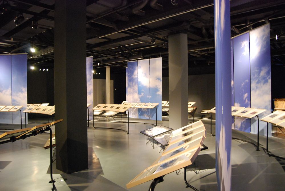 Die Ausstellungsansicht von "Eduard Spelterini Fotografien des Ballonpioniers" zeigt die Aneinanderreihung von Bilderrahmen, die im Raum angeordnet sind. Im Hintergrund stehen Banner, die mit einem Himmel und Wolken Motiv bedruckt sind.