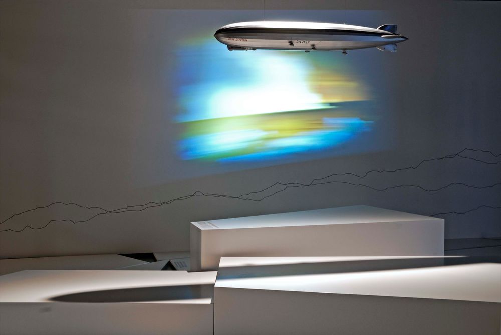 Die Ausstellungsansicht zu "66 Grad 30 Minuten Nord. Luftschiffe über der Arktis" zeigt ein von der Decke hängendes Luftschiffmodell, dahinter ist eine verschwommene Landschaft an die Wand projiziert.