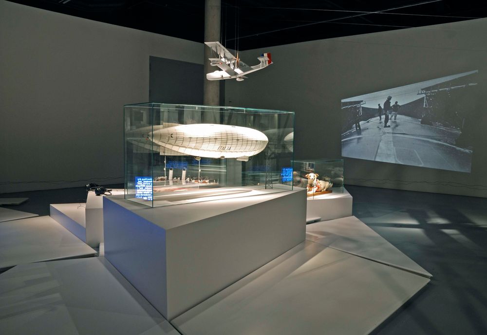 Die Ausstellungsansicht zu "66 Grad 30 Minuten Nord. Luftschiffe über der Arktis" zeigt zwei Vitrinen, die auf weißen Fächen stehen. Über den Vitrinen hängt ein Flugzeugmodell.
