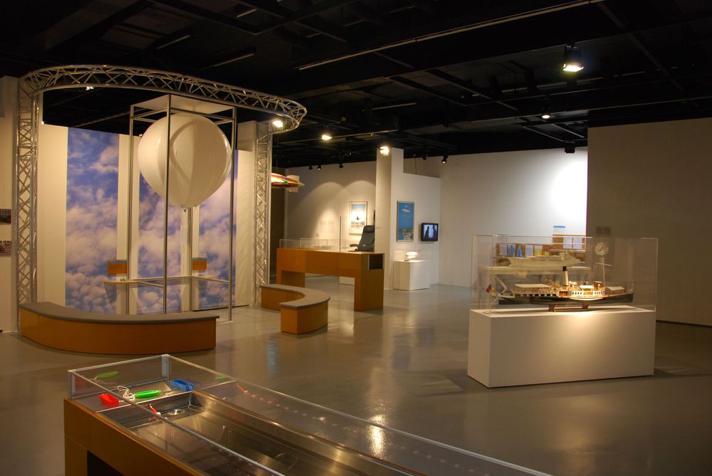Die Ausstellungsansicht von "Wasser, Straße, Schiene, Luft. Mobilität am Bodensee" zeigt mehrere Experimentierstationen und Vitrinen, in denen Exponate ausgestellt sind. 