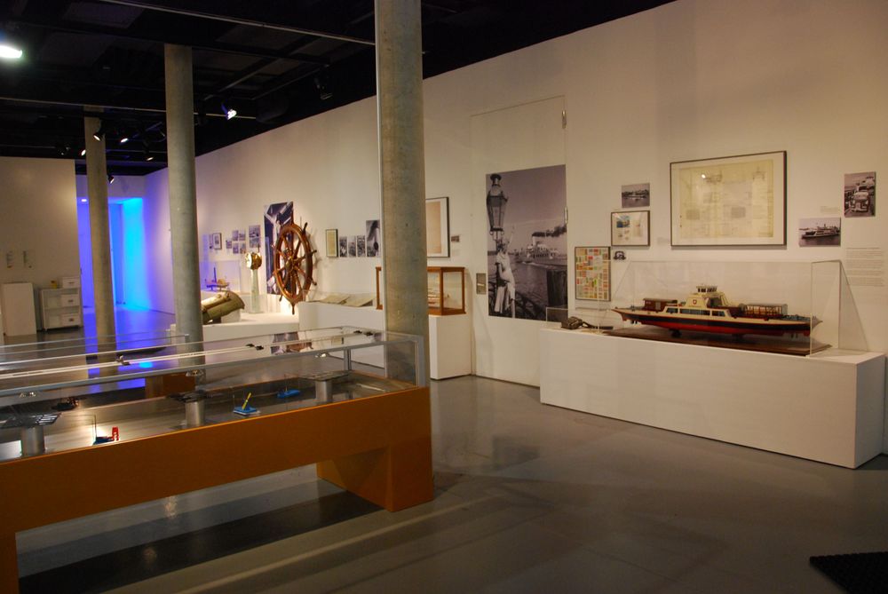 Die Ausstellungsansicht von "Wasser, Straße, Schiene, Luft. Mobilität am Bodensee" zeigt mehrere Experimentierstationen und Vitrinen, in denen Exponate ausgestellt sind. 