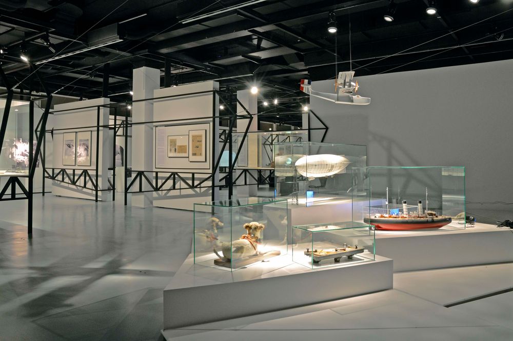 Die Ausstellungsansicht zu "66 Grad 30 Minuten Nord. Luftschiffe über der Arktis" zeigt Vitrinen, die auf weißen Fächen stehen. Über den Vitrinen hängt ein Flugzeugmodell.