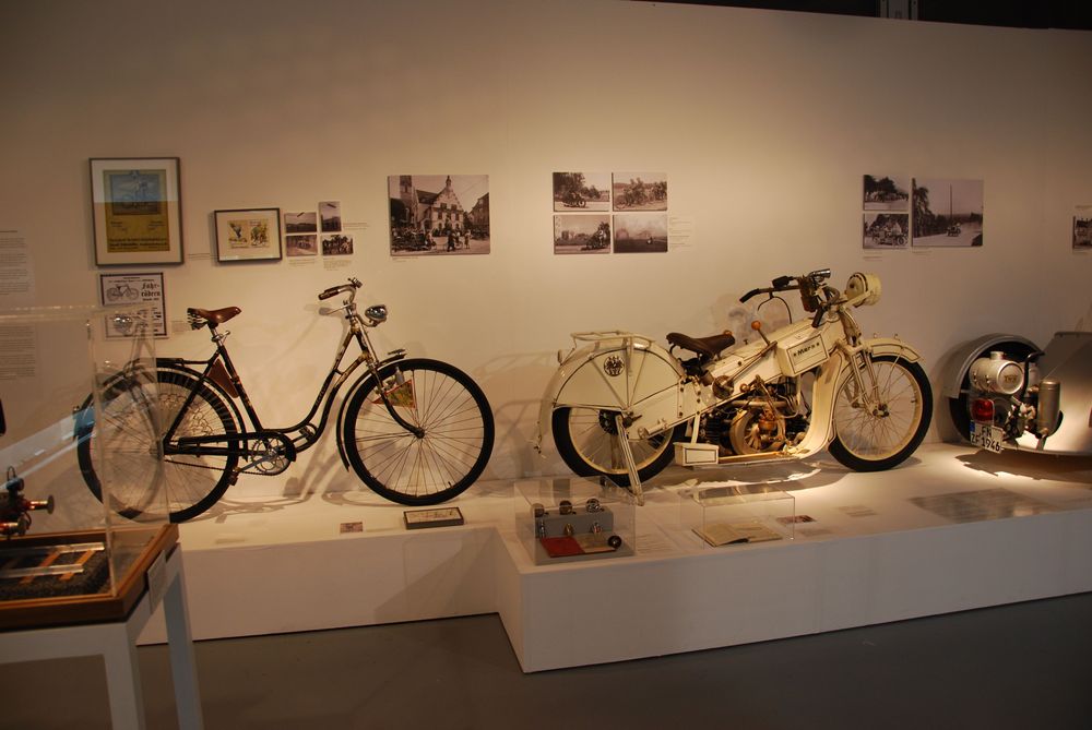 Die Ausstellungsansicht von "Wasser, Straße, Schiene, Luft. Mobilität am Bodensee" zeigt ein Podest, auf dem ein altes Fahrrad und ein altes Moped stehen. An der Wand dahinter hängen mehrere Bilder.