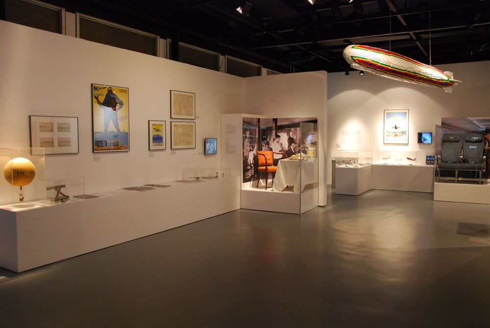 Die Ausstellungsansicht von "Wasser, Straße, Schiene, Luft. Mobilität am Bodensee" zeigt mehrere Schaukästen und Vitrinen, in denen Exponate ausgestellt sind. Ein aus LEGO-Bausteinen bestehender Zeppelin hängt von der Decke. 
