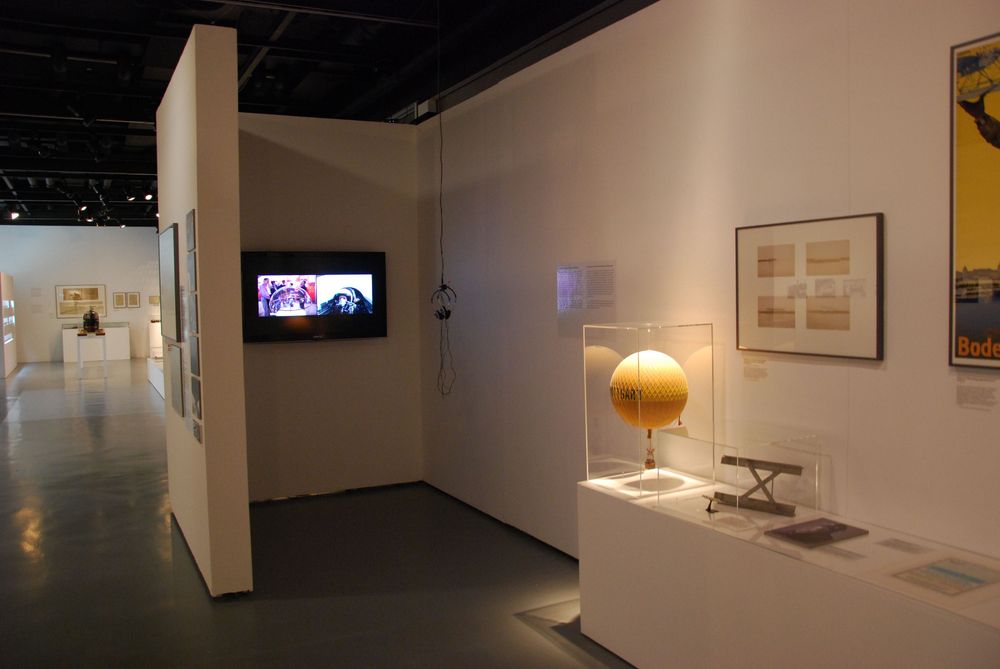 Die Ausstellungsansicht von "Wasser, Straße, Schiene, Luft. Mobilität am Bodensee" zeigt eine Videostation, bei der Kopfhörer von der Decke hängen. Rechts daneben befinden sich Vitrinen mit Exponaten.