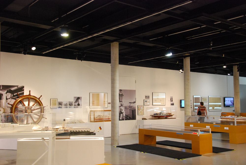 Die Ausstellungsansicht von "Wasser, Straße, Schiene, Luft. Mobilität am Bodensee" zeigt mehrere Vitrinen und Experimentierstationen. Eine Frau betrachtet die Bilder, die an der Wand hängen.