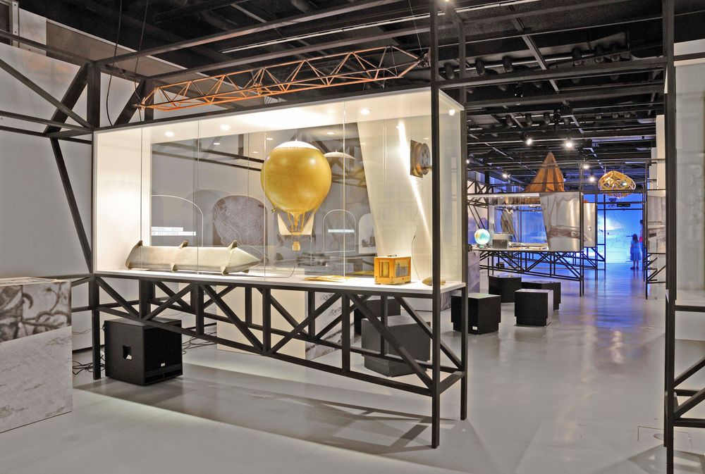 Die Ausstellungsansicht zu "66 Grad 30 Minuten Nord. Luftschiffe über der Arktis" zeigt mehrere größere Vitrinen mit Exponaten und quadratischen Sitzmöglichkeiten.