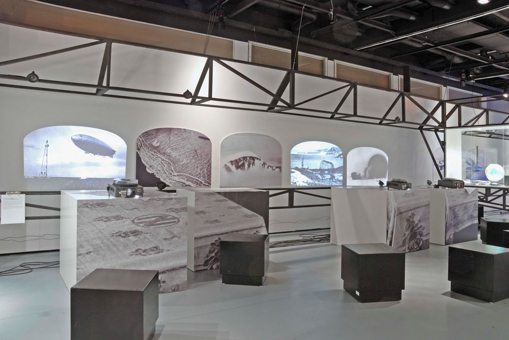 Die Ausstellungsansicht zu "66 Grad 30 Minuten Nord. Luftschiffe über der Arktis" zeigt mehrere Beamer, die historische Aufnahmen an die Wand spielen. Im Raum befinden sich quadratische Sitzmöglichkeiten.