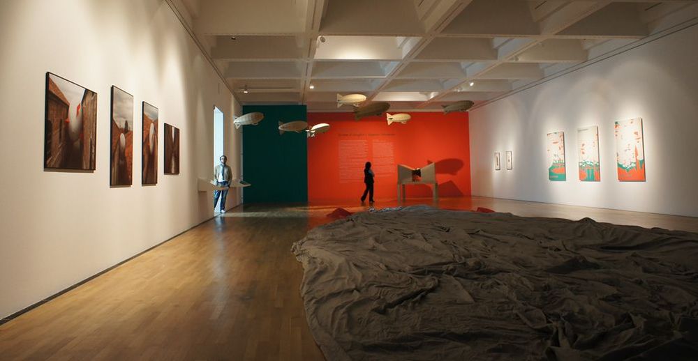 Die Ausstellungsansicht von "Héctor Zamora Sciame di dirigibili / Zeppelin-Schwärme" zeigt einen fast leeren Ausstellungsraum in dem ein großes Tuch auf dem Boden liegt. An den Wänden hängen verschiedene Bilder.
