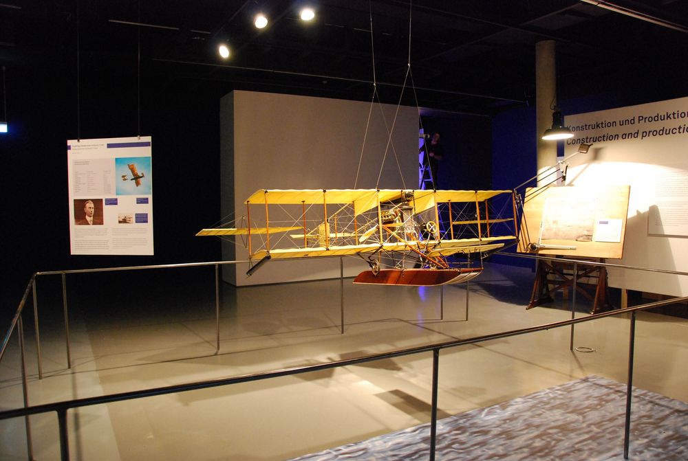 In der Mitte des Raumes hängt ein Flugzeugmodell von der Decke. Rechts und links dahinter hängen Ausstellungstexte und Bilder.