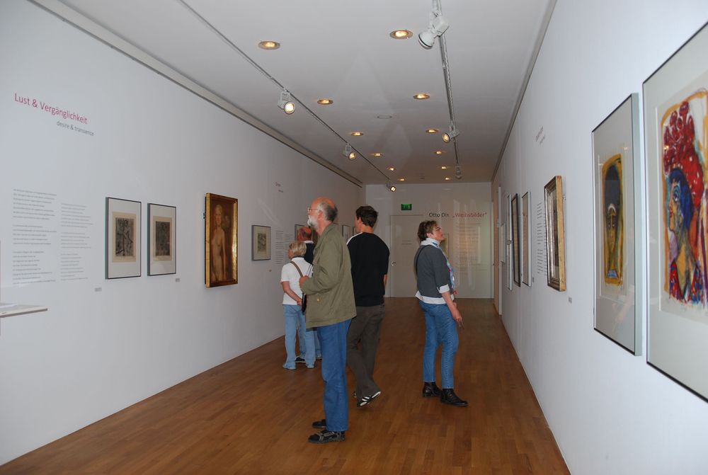 Die Ausstellungsansicht von "Otto Dix - Weibsbilder" zeigt einen Flur, an dessen Wänden unterschiedliche Gemälde hängen. Eine Gruppe von Menschen betrachtet die Gemälde. 