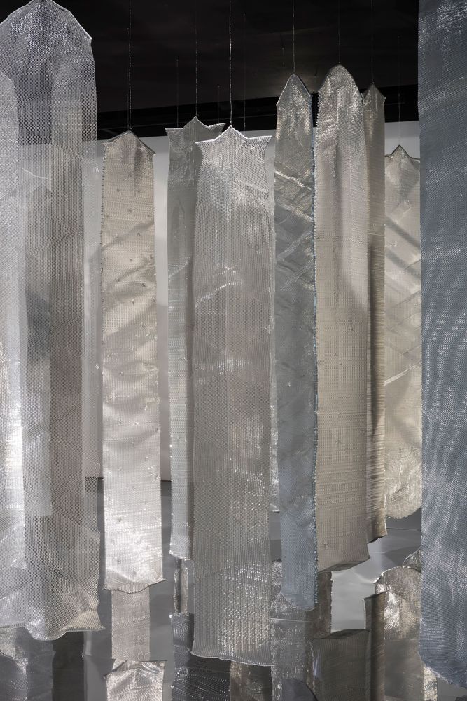 Eine Installation aus mehreren metallisch glänzenden Stoffen hängt von der Decke des Raumes. 